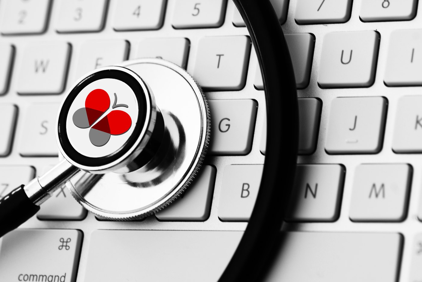 A stethoscope lying across a white Mac keyboard, photographed close in high-contrast black and white. The Visova butterfly mark sits inside the stethoscope's diaphragm, where the membrane would be, and its red is the only color anywhere in the frame.